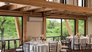 A dining area with tables set for a meal, large windows offering a view of greenery, and wooden beams on the ceiling.