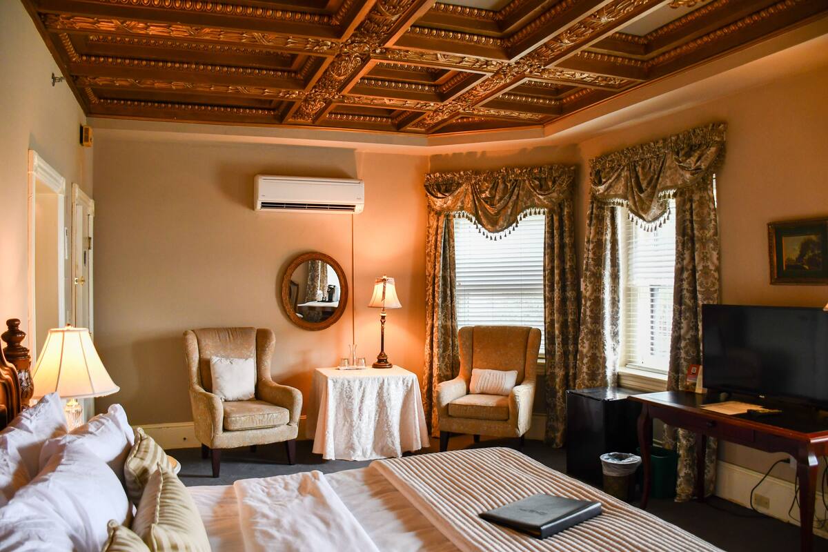 A hotel room with a bed, two armchairs, a small table with a lamp, a television, and a ceiling with intricate woodwork.