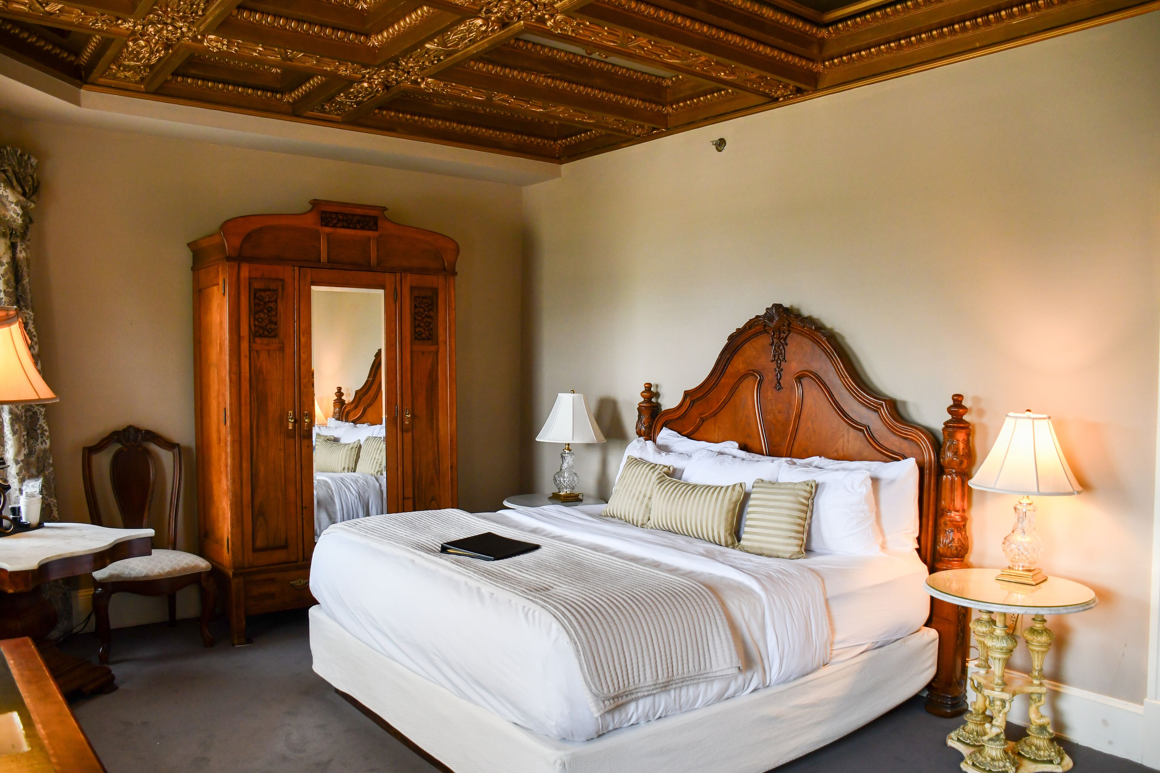 A large bed with a wooden headboard, two bedside lamps, a nightstand, a wooden wardrobe, and a desk with a chair.