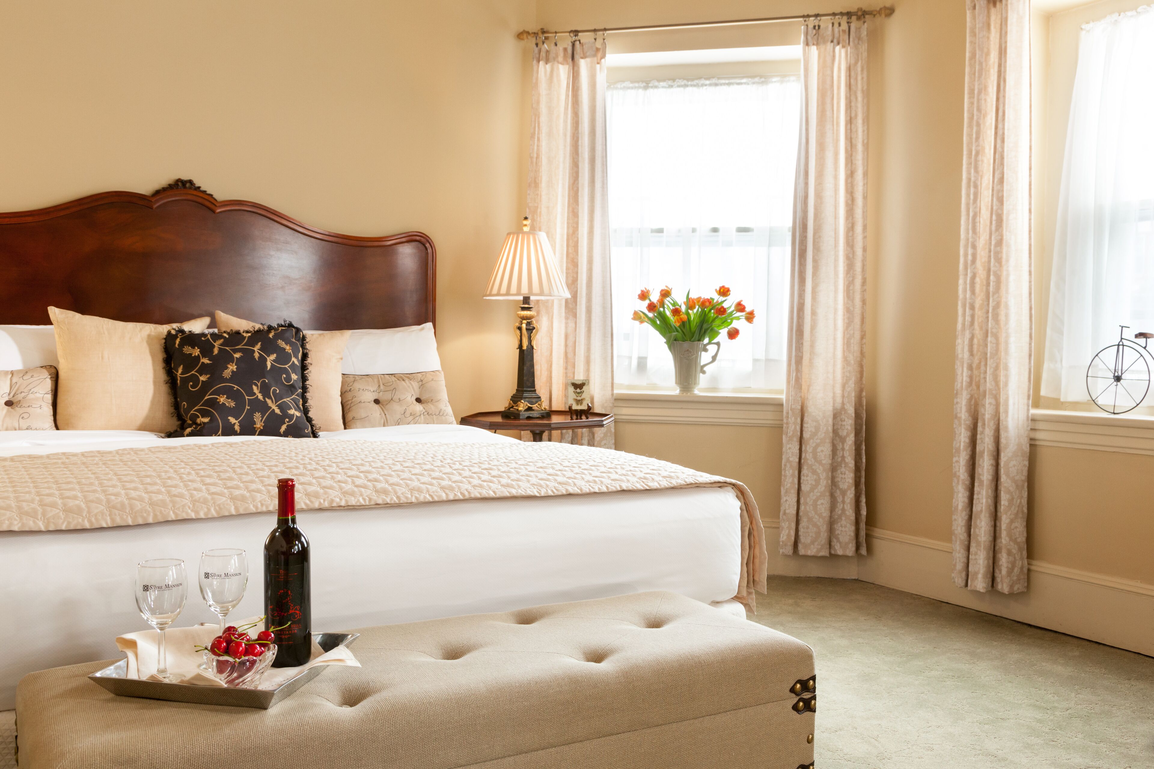 Een hotelkamer met een groot bed, een nachtkastje met een lamp, een raam met gordijnen, en een dienblad met een fles en glazen.