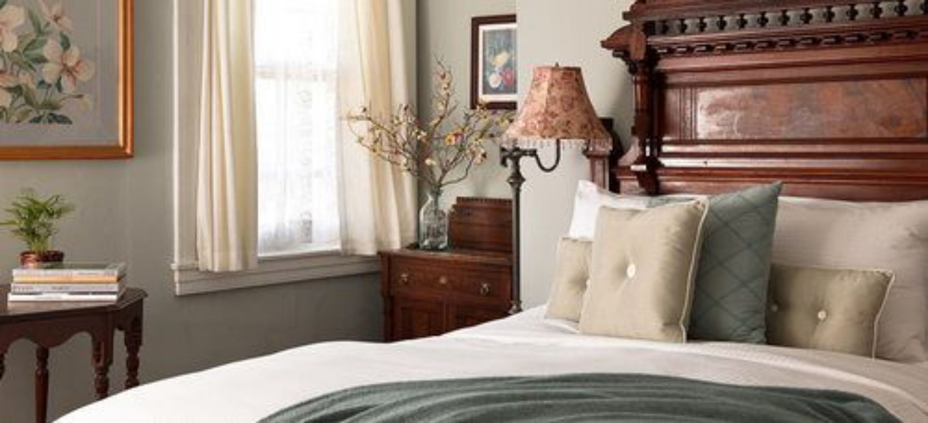 A bedroom with a large bed, a nightstand with a lamp, a dresser, and a window with curtains.