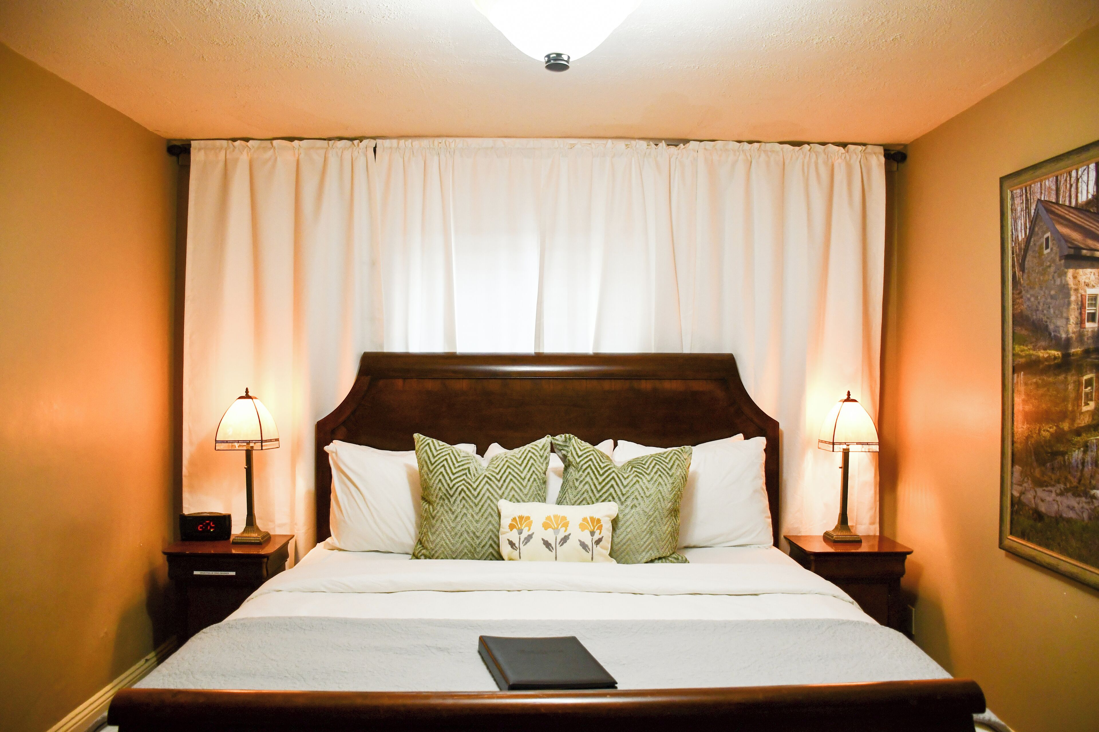 A neatly made bed with a white comforter and green patterned pillows, flanked by two bedside tables with lamps, in a room with cream-colored walls and a wooden headboard.