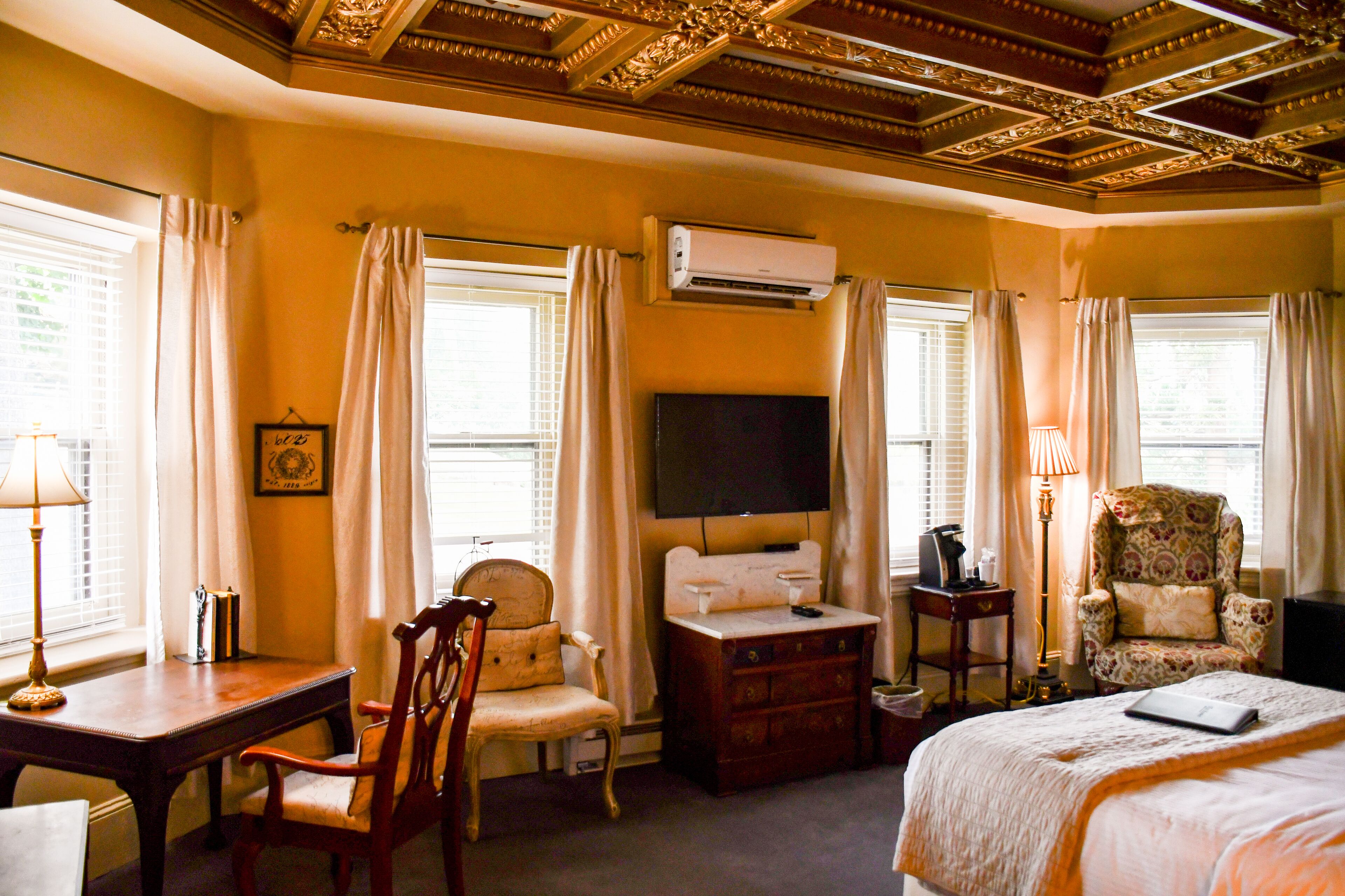 Una habitación de hotel con un escritorio de madera, una silla, una cama con edredón blanco, un televisor y un equipo de aire acondicionado.