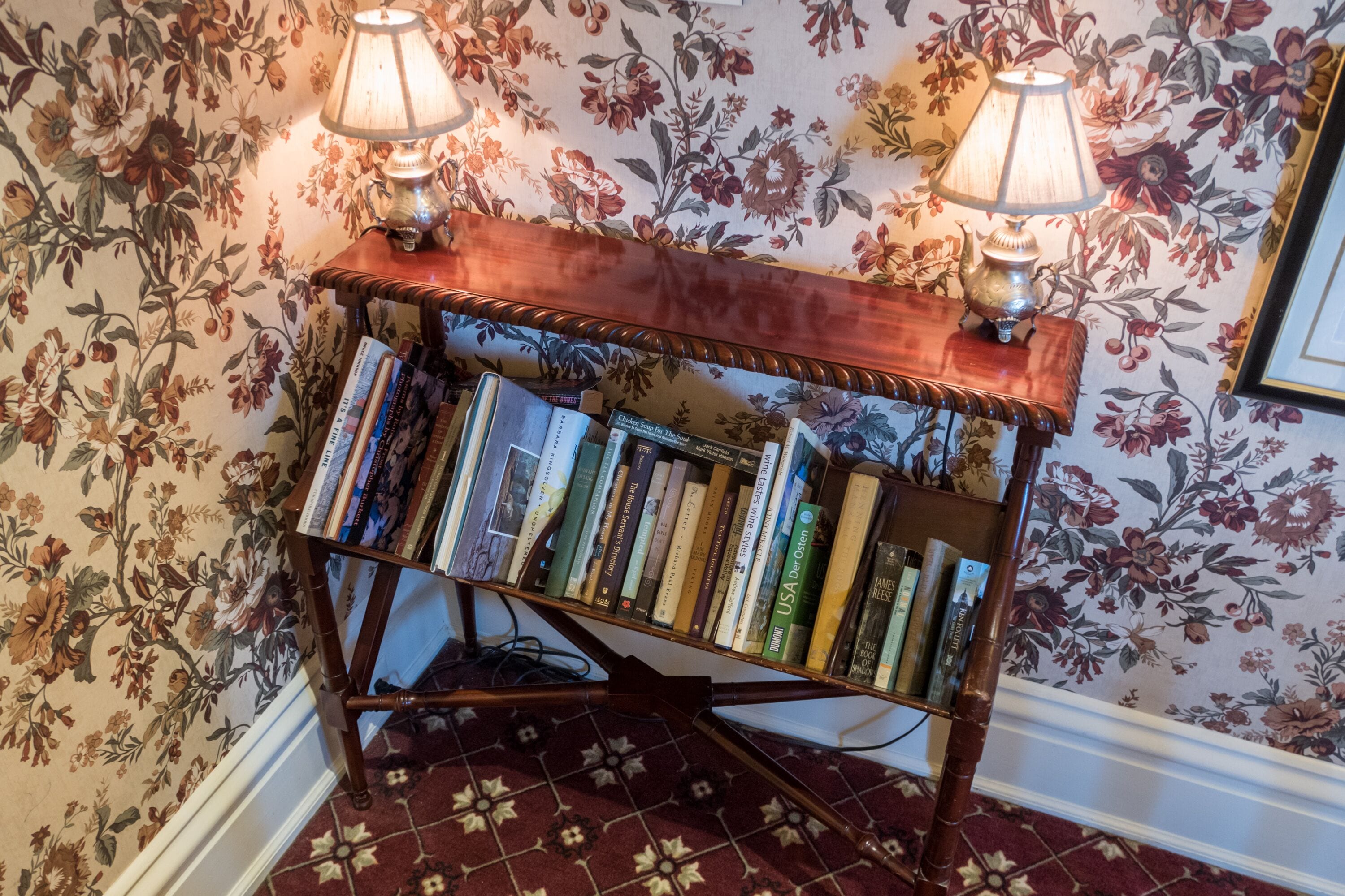Une bibliothèque en bois, ornée de livres et de deux lampes, placée devant un mur recouvert d’un papier peint à motifs floraux.
