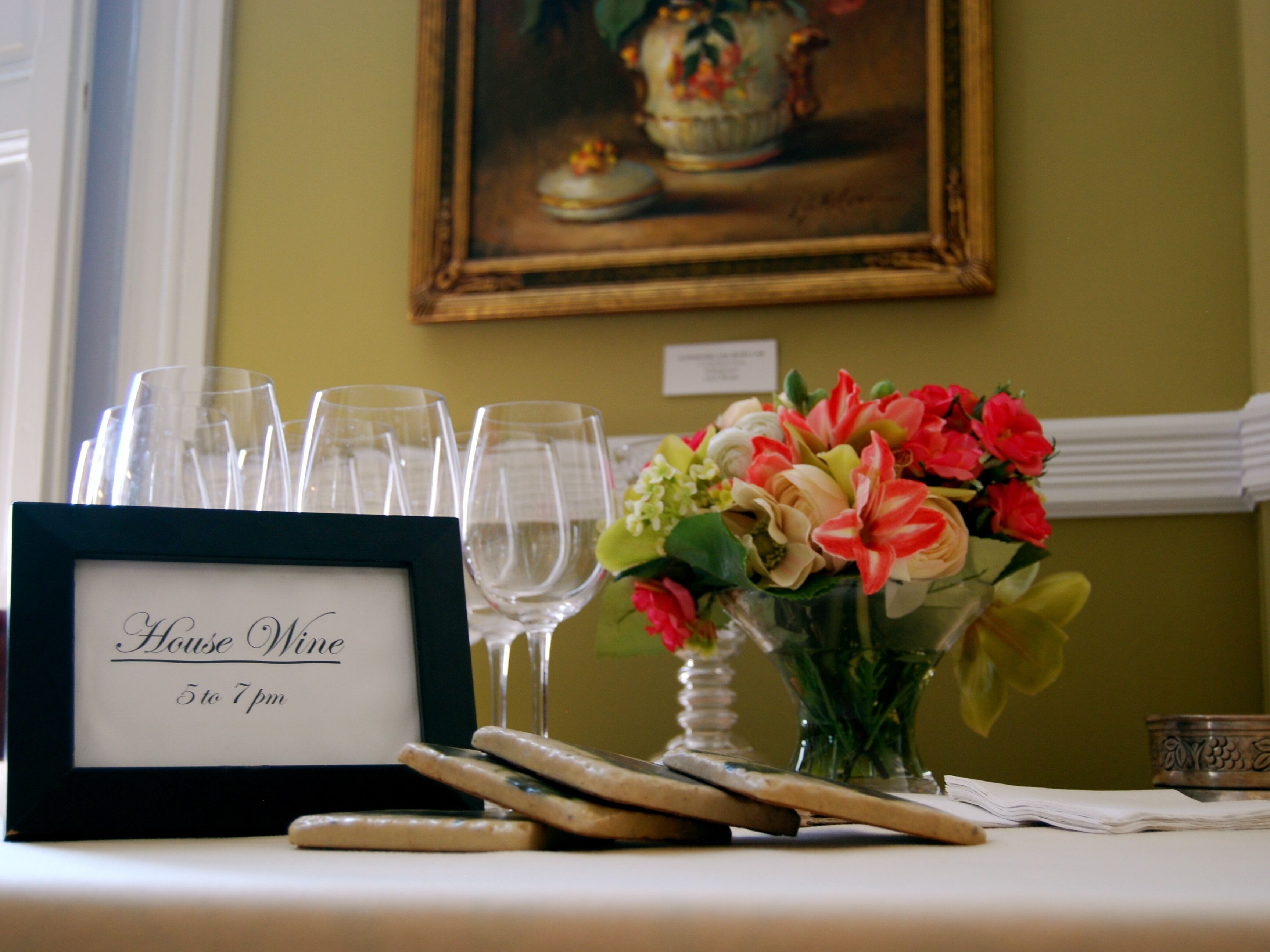 A table with a framed sign reading "House Wine 5 to 7 pm", wine glasses, a vase with flowers, and a painting on the wall.