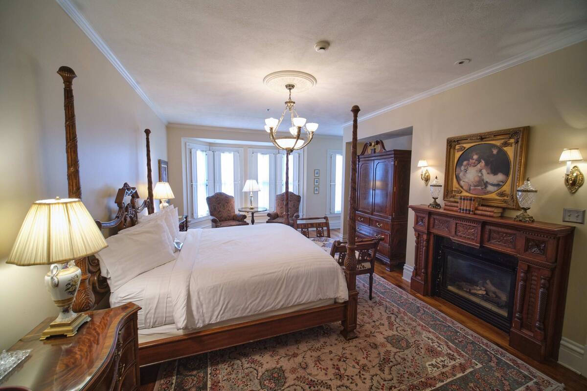 A traditional bedroom with a four-poster bed, a fireplace, and a large window.