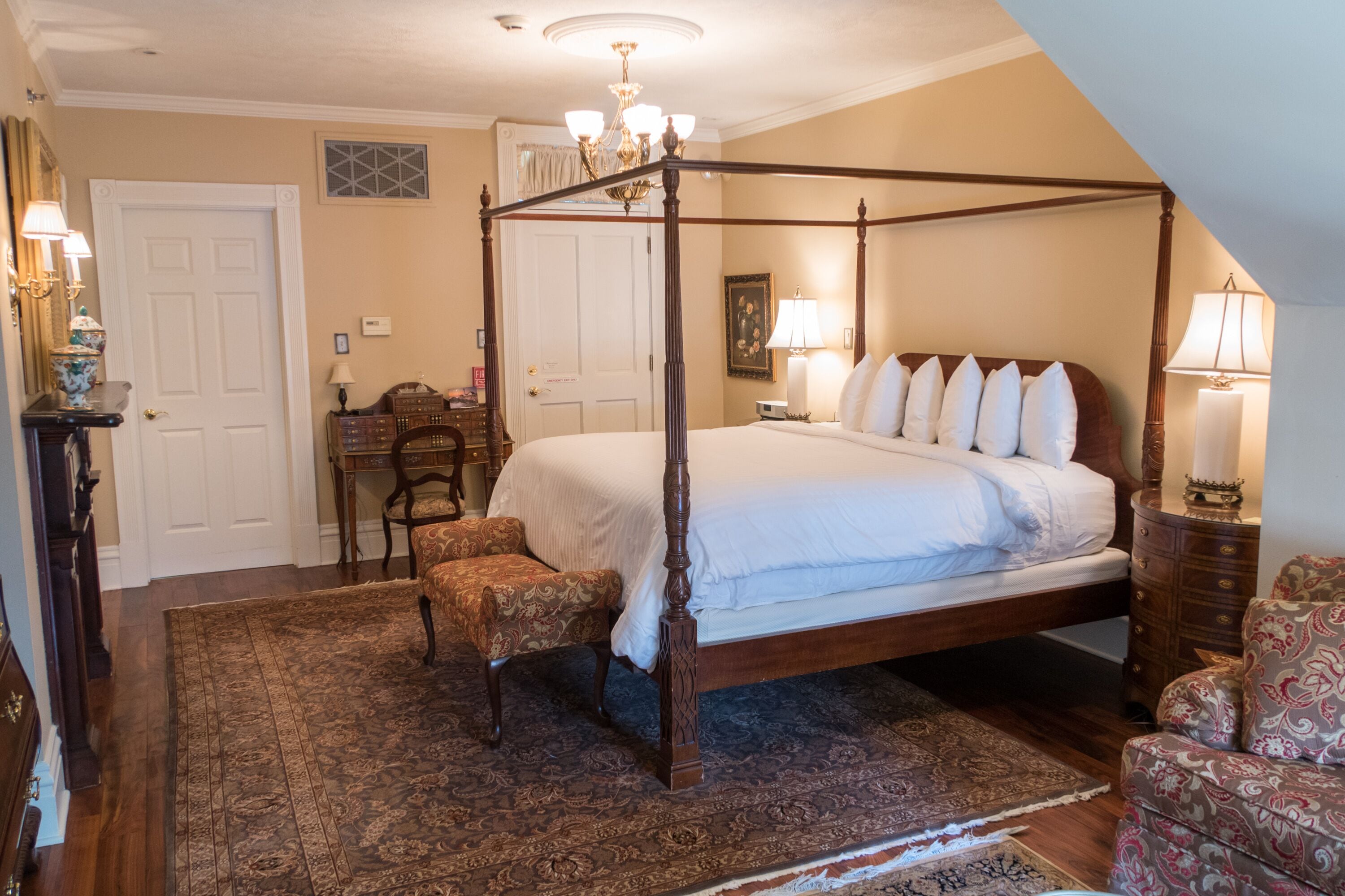 A four-poster bed with a wooden frame, a matching nightstand, a chair, and a patterned rug in a room with a sloped ceiling and a chandelier.