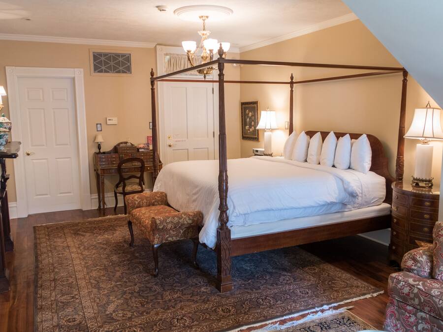 A four-poster bed with a wooden frame, a matching nightstand, a chair, and a patterned rug in a room with a sloped ceiling and a chandelier.