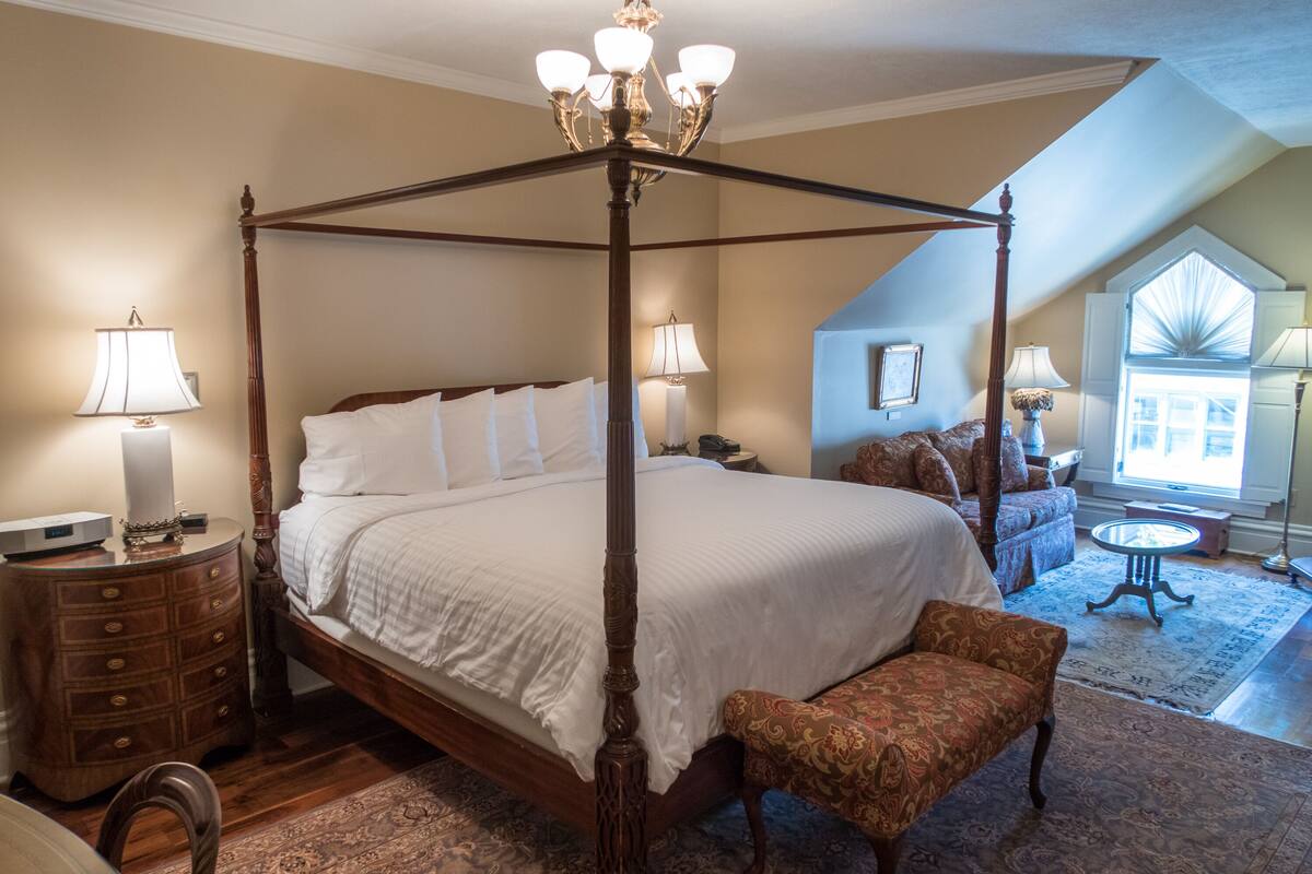 A four-poster bed with a wooden frame and white bedding, a wooden nightstand with drawers, a patterned armchair, and a small round table with a lamp.