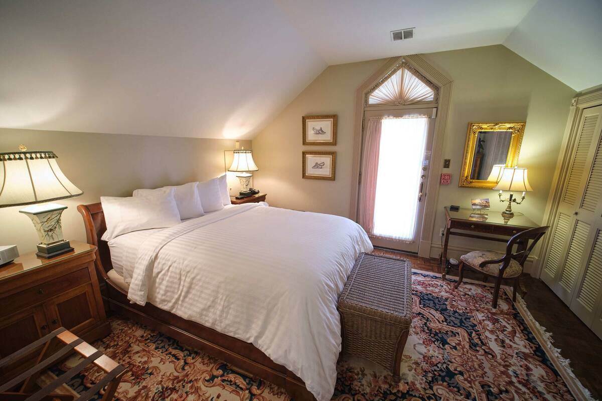 A bedroom with a large bed, two bedside tables with lamps, a chair, a desk with a lamp, a mirror, and a window with curtains.