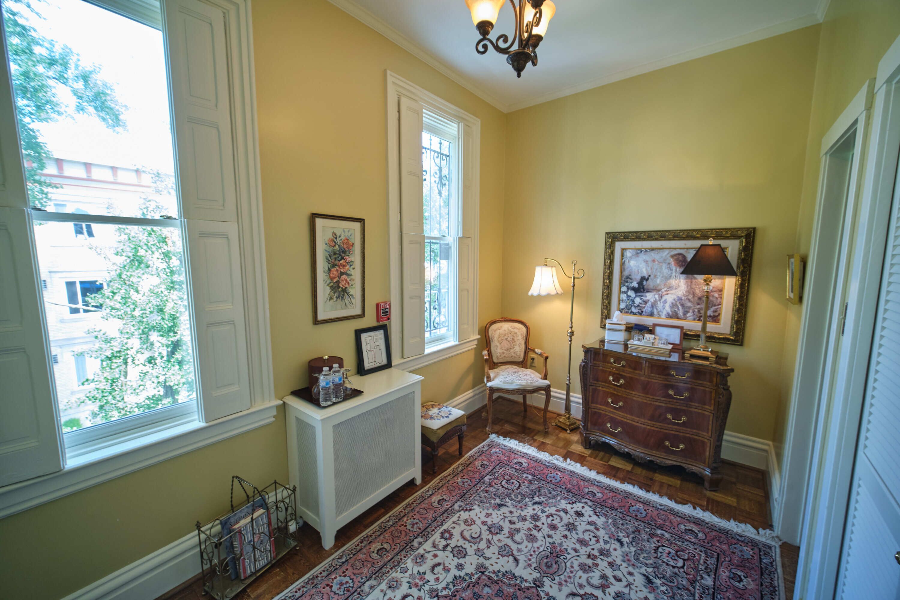 A room with a large window, a radiator, a chair, a dresser, and a rug.