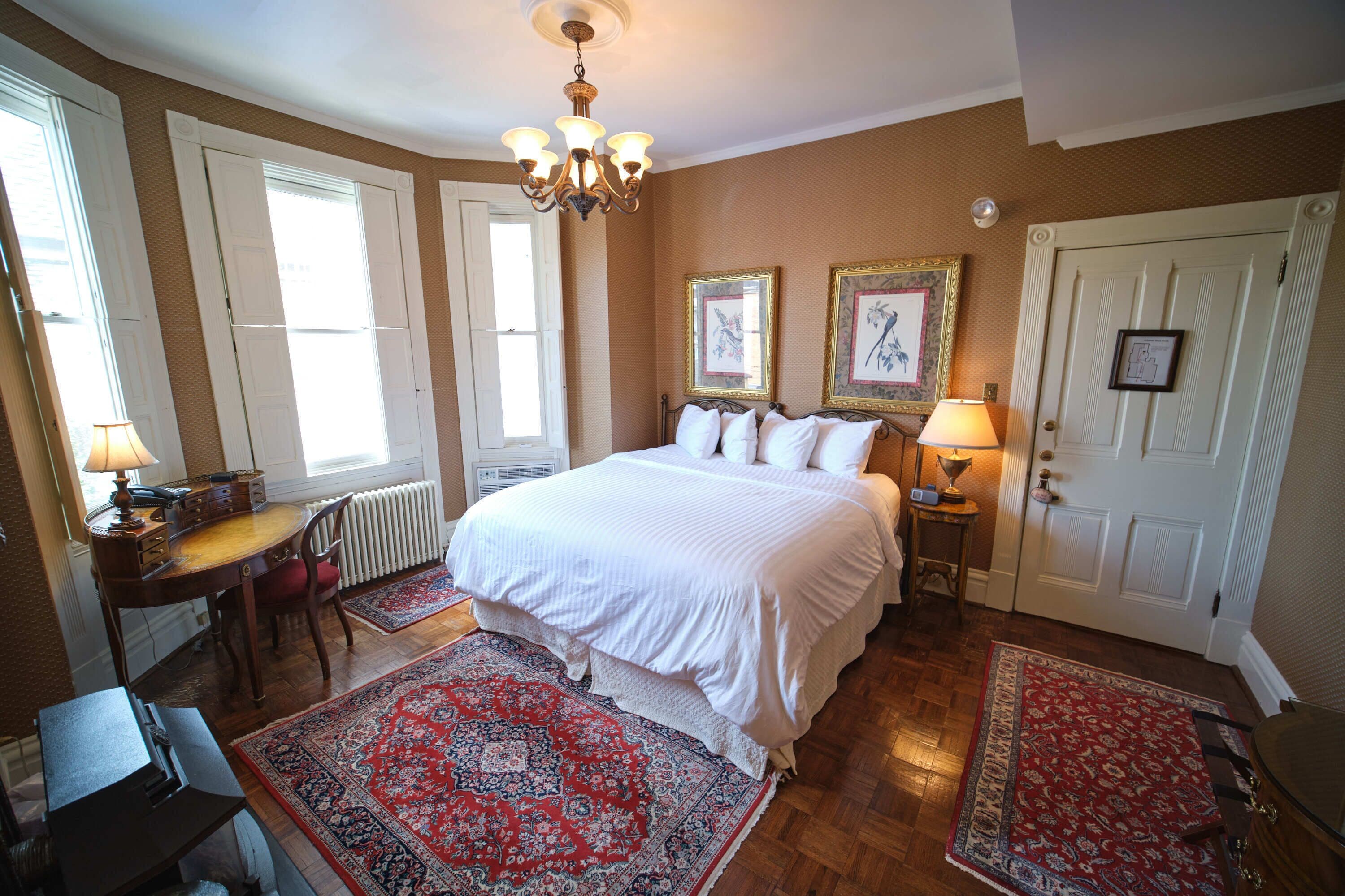 A bedroom with a large bed, a desk with a lamp, a chair, two framed pictures, and a red and white patterned rug.