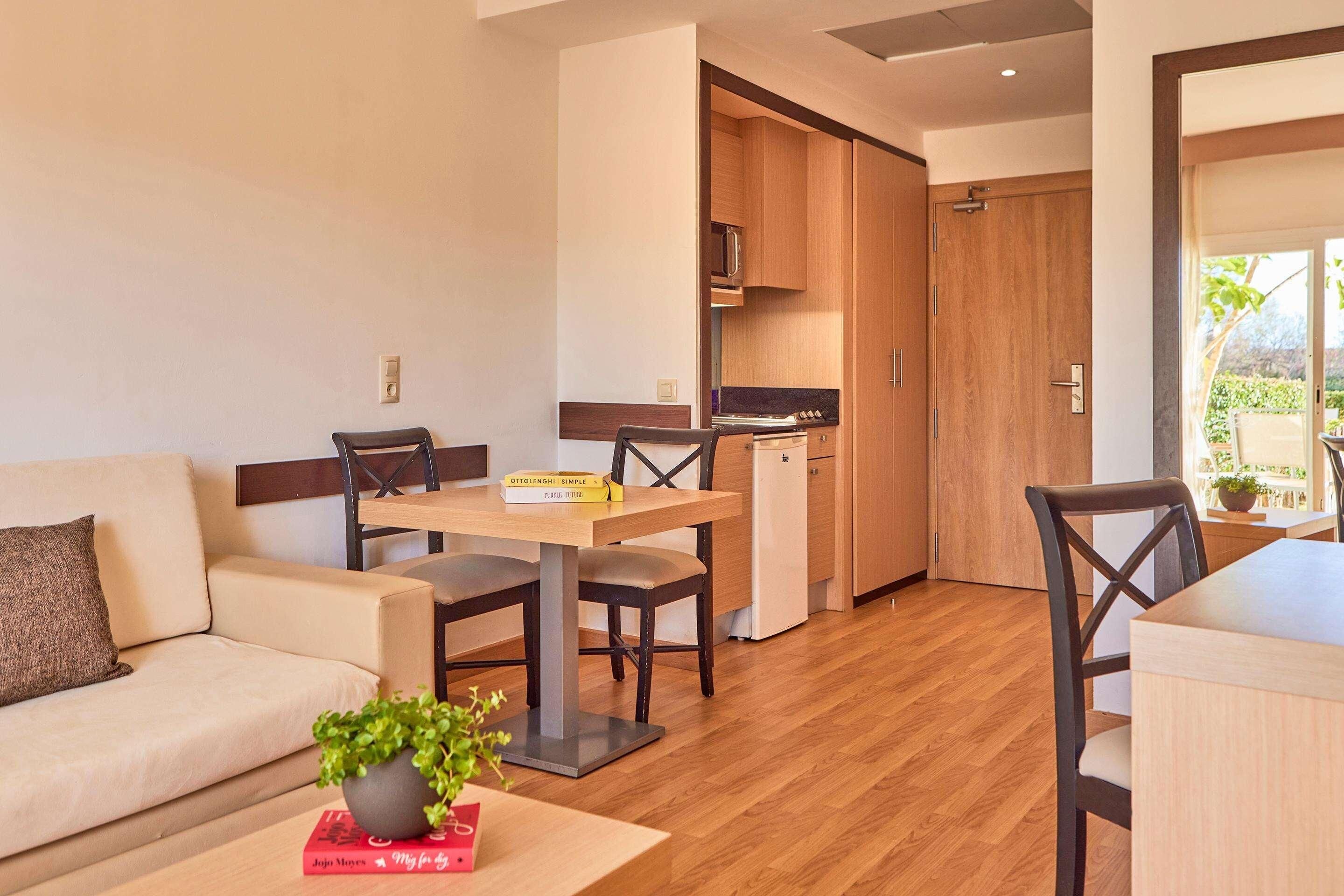 A modern living room with a sofa, a dining table with chairs, a small kitchen area, and a sliding door leading to an outdoor space.