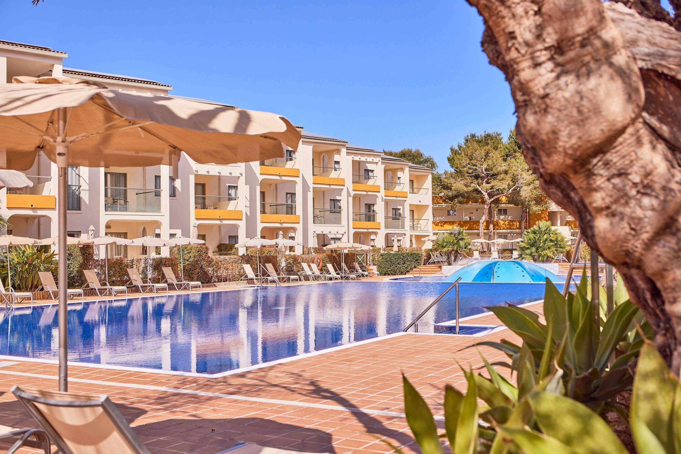 Área de piscina com espreguiçadeiras e guarda-sóis, cercada por um edifício com varandas.