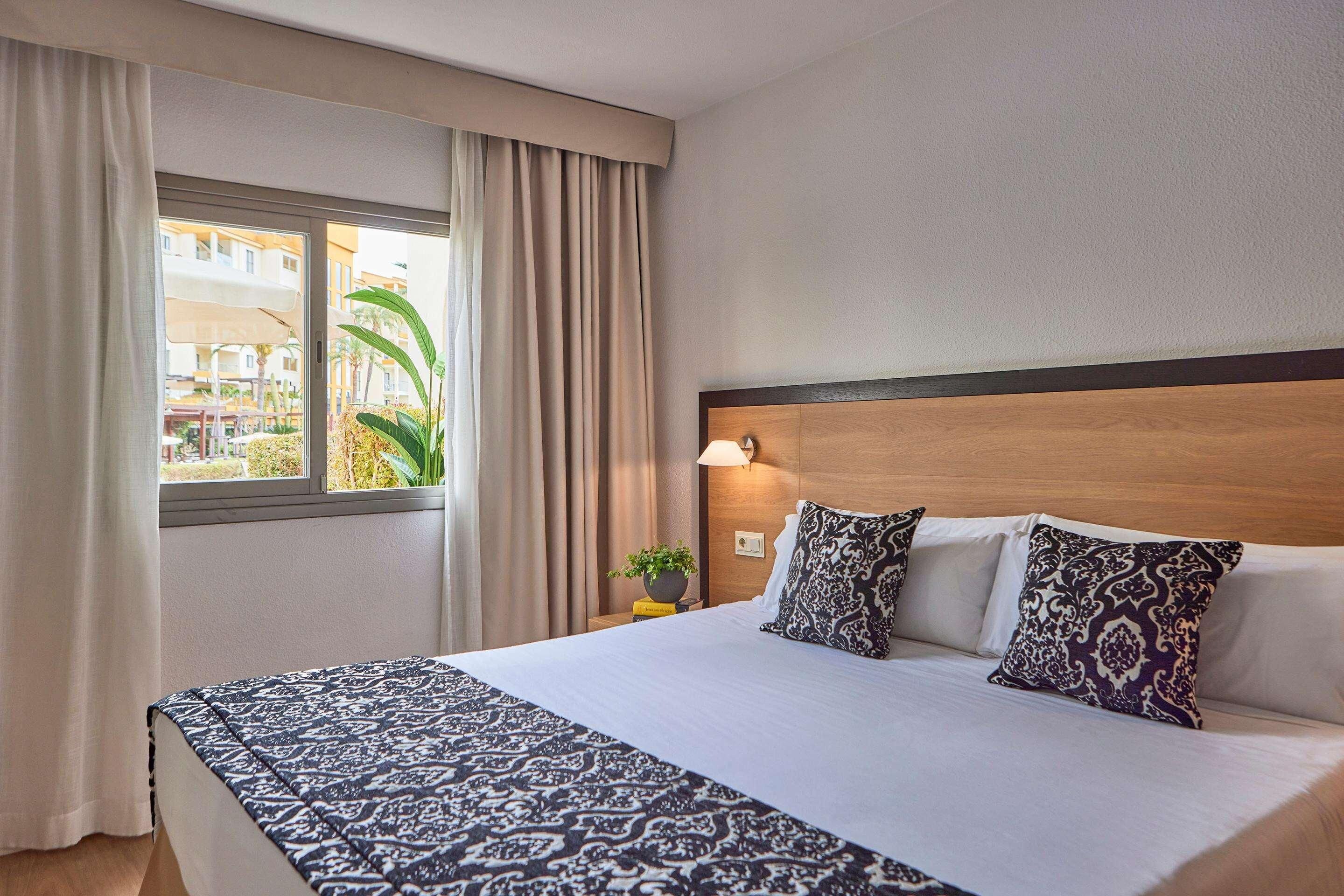 Habitación de hotel con una cama grande, cabecera de madera, mesita de noche con lámpara y ventana con cortinas.