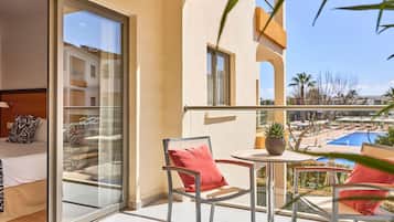 A balcony with a glass door, a bed with patterned pillows, a small table with a plant, and a view of a swimming pool and buildings.