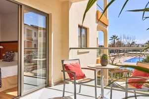 Un balcón con puerta de cristal, una cama con almohadas estampadas, una mesita con una planta y vistas a una piscina y edificios.