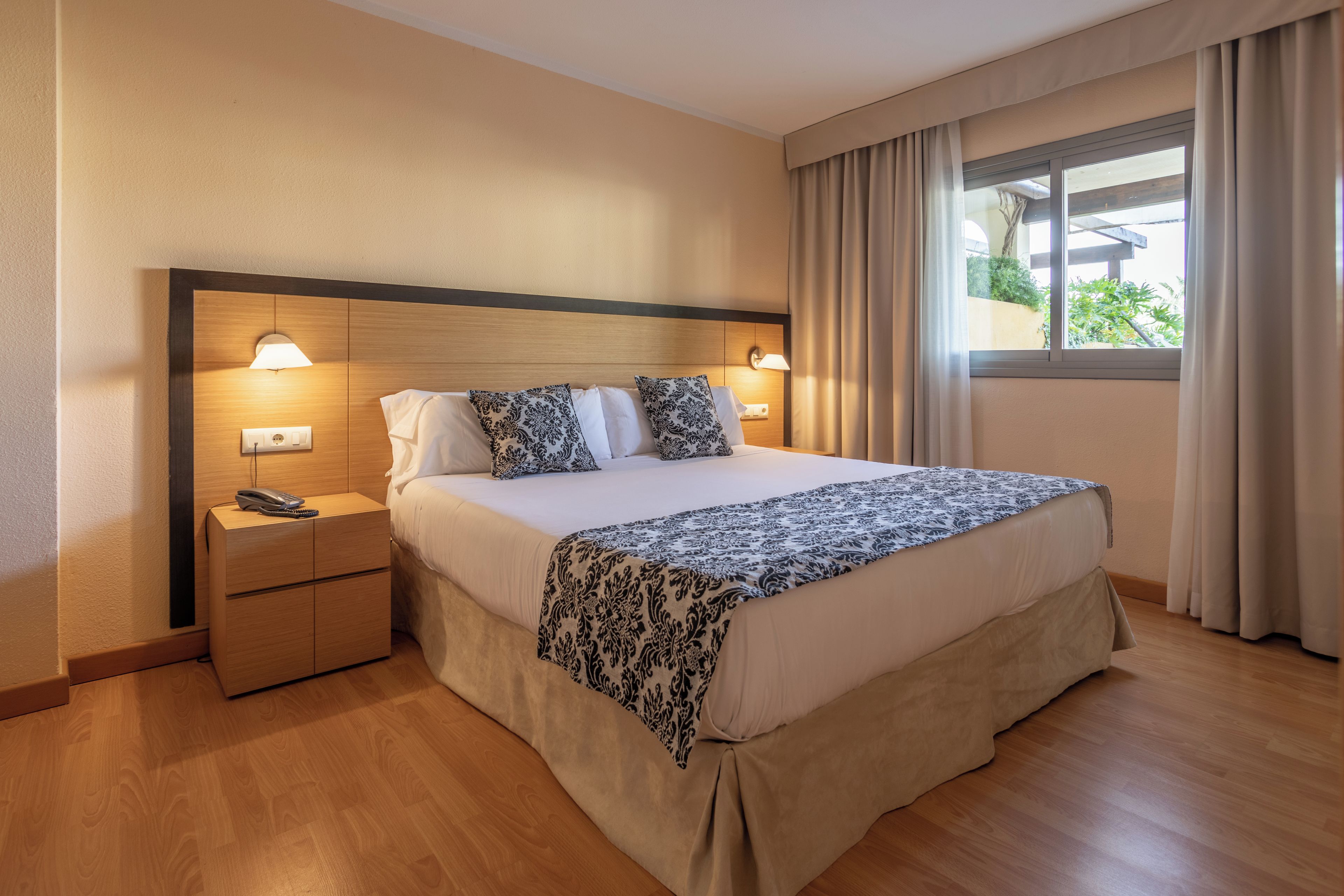 Ein Hotelzimmer mit einem großen Bett, Nachttischlampen, einem Holznachttisch und einem Fenster mit Vorhängen.