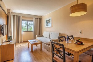 Habitación de hotel moderna con una mesa de comedor de madera, un sofá, una pequeña mesa de centro, un televisor y una ventana con cortinas.