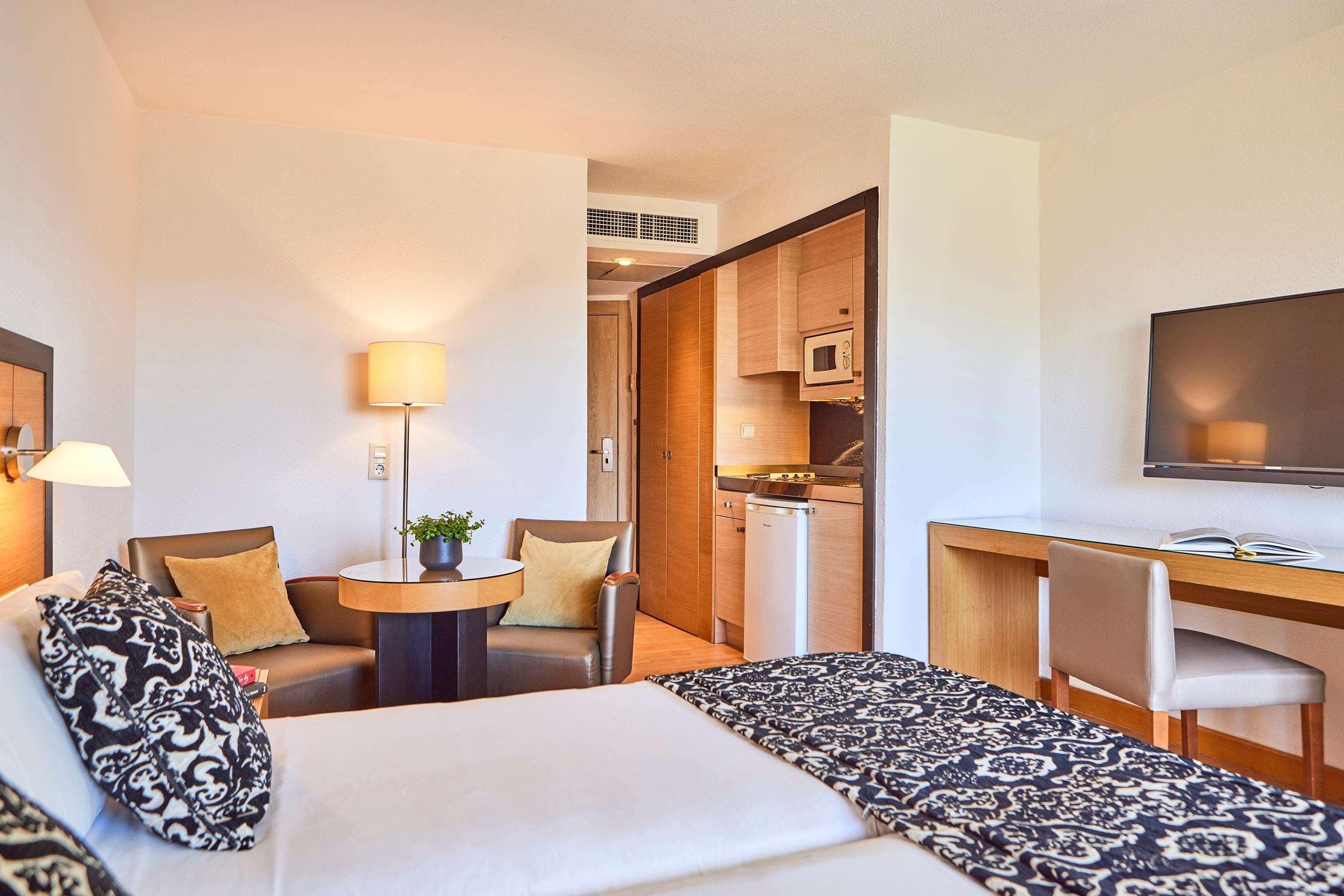 Habitación de hotel con cama, sofá, escritorio, silla, televisor, lámpara y kitchenette.