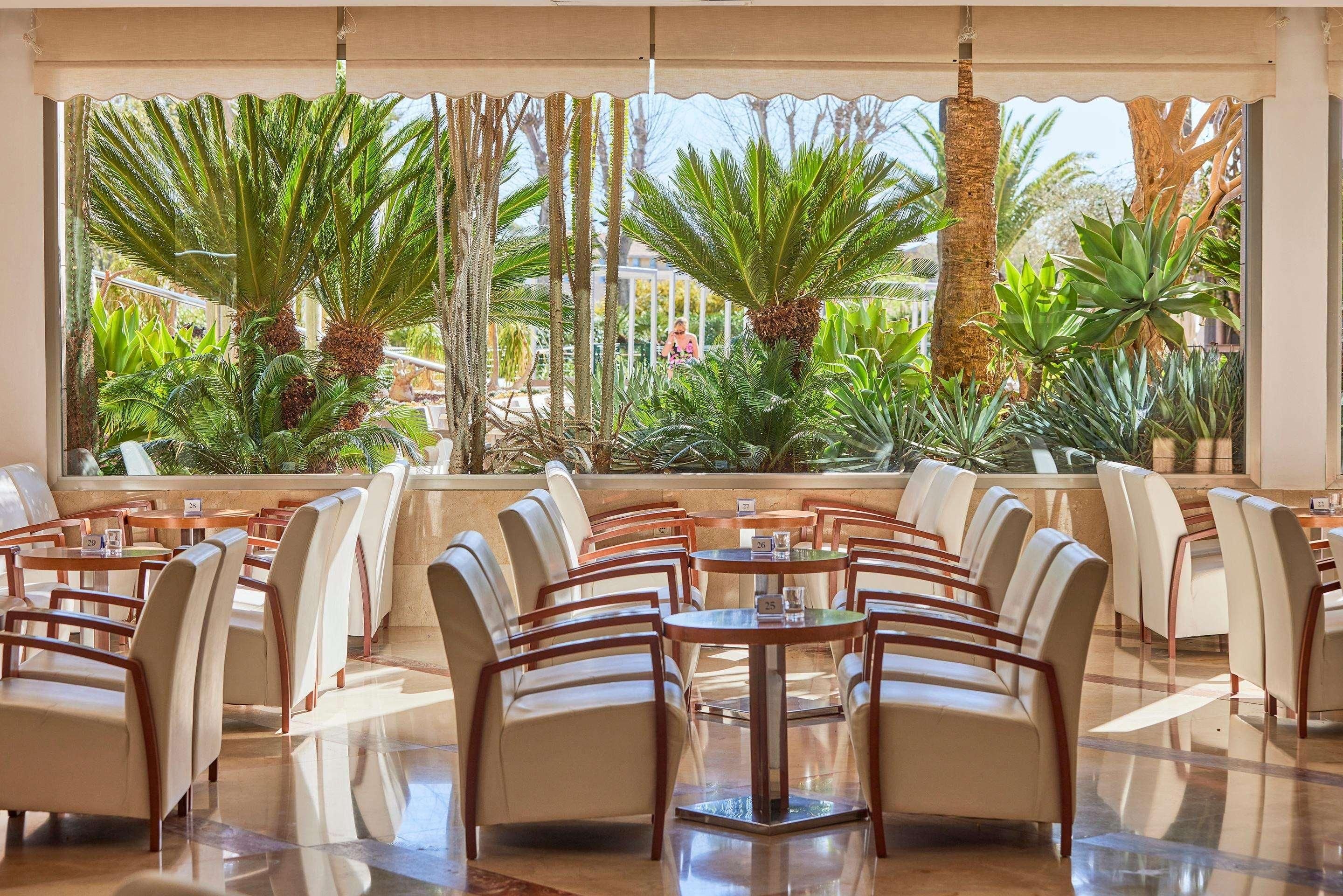 Een restaurant met uitzicht op tropische planten en palmbomen. Tafels en stoelen staan er klaar voor een maaltijd.