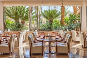 Un restaurante con vistas a plantas tropicales y palmeras. Mesas y sillas dispuestas para comer.