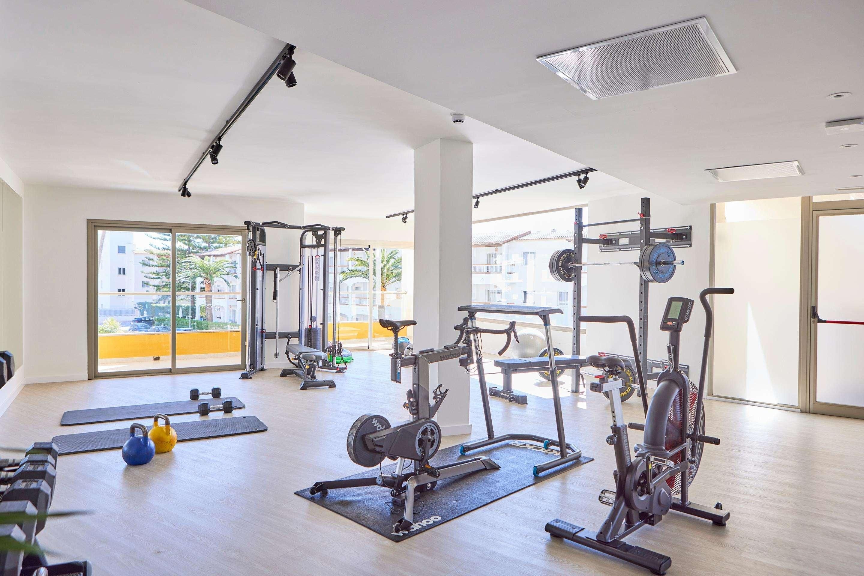 Un gimnasio con diversos equipos de ejercicio, ventanas amplias y vistas a edificios exteriores.