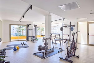 Un gimnasio con diversos equipos de ejercicio, ventanas amplias y vistas a edificios exteriores.
