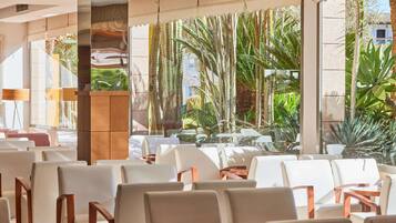 Un vestíbulo de hotel con filas de sillas blancas, un mostrador de recepción de madera y grandes ventanales que ofrecen vistas a plantas tropicales y un edificio exterior.