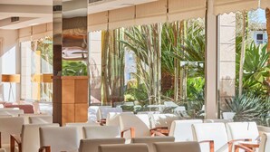 Un vest íbulo de hotel con filas de sillas blancas, un mostrador de recepción de madera y grandes ventanales que ofrecen vistas a plantas tropicales y un edificio exterior.
