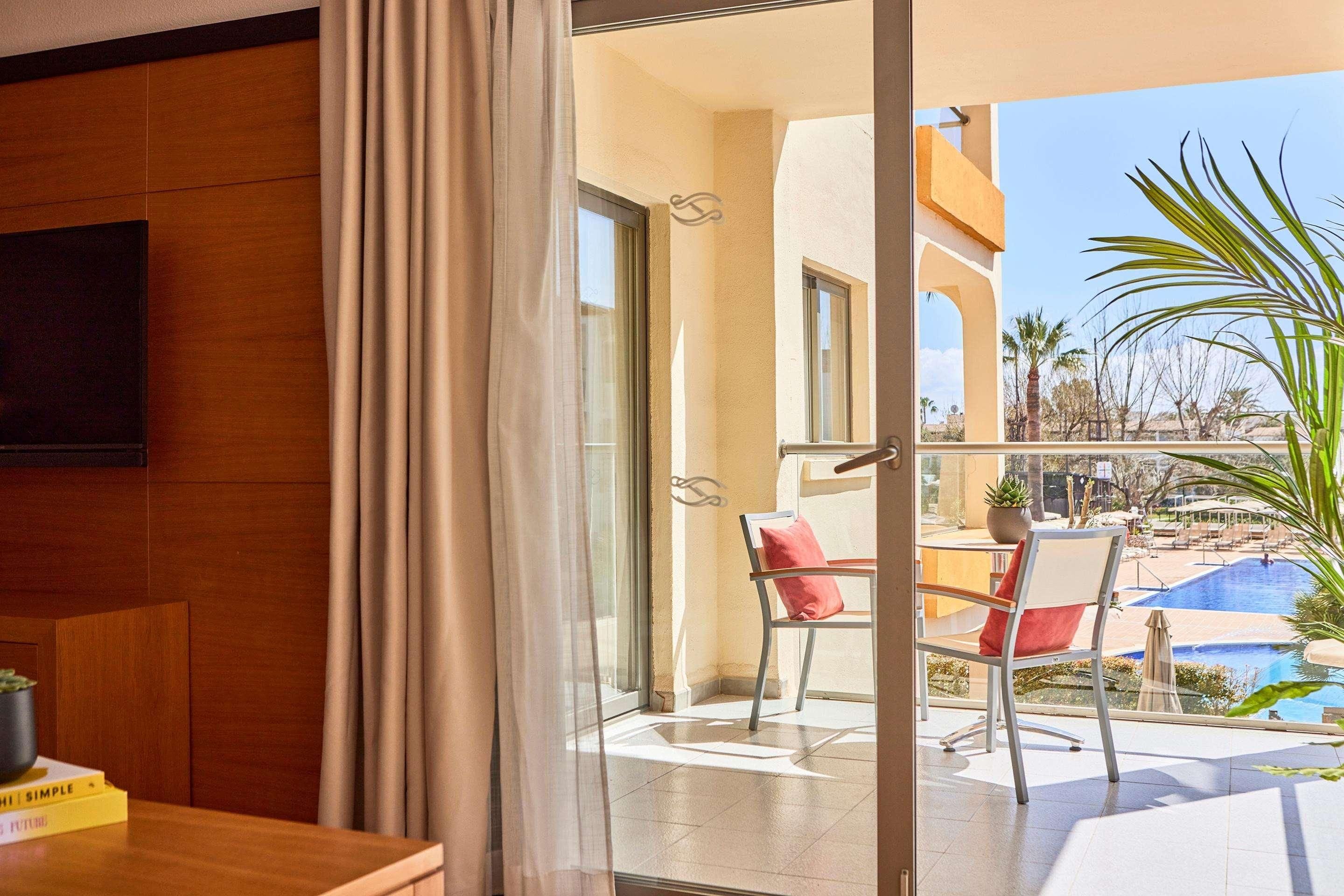 Een hotelkamer met balkon, een eettafel en een stoel met een rode kussen.