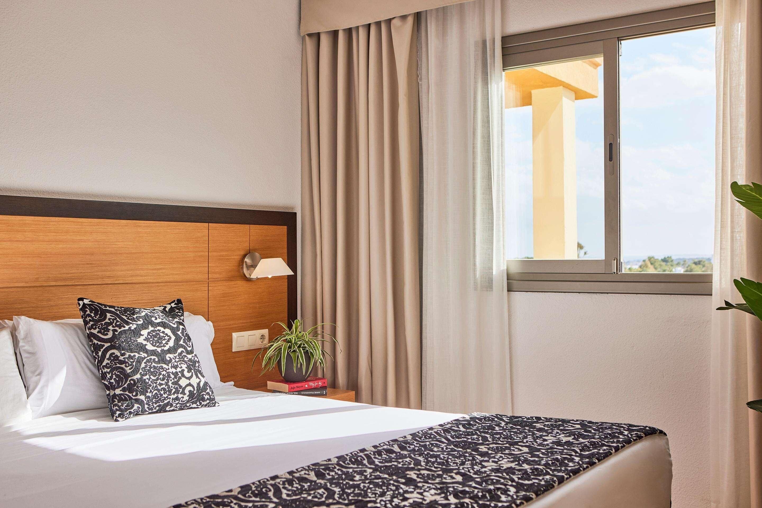 Una habitación de hotel con cama, mesita de noche, lámpara, ventana con cortinas y vistas al exterior.