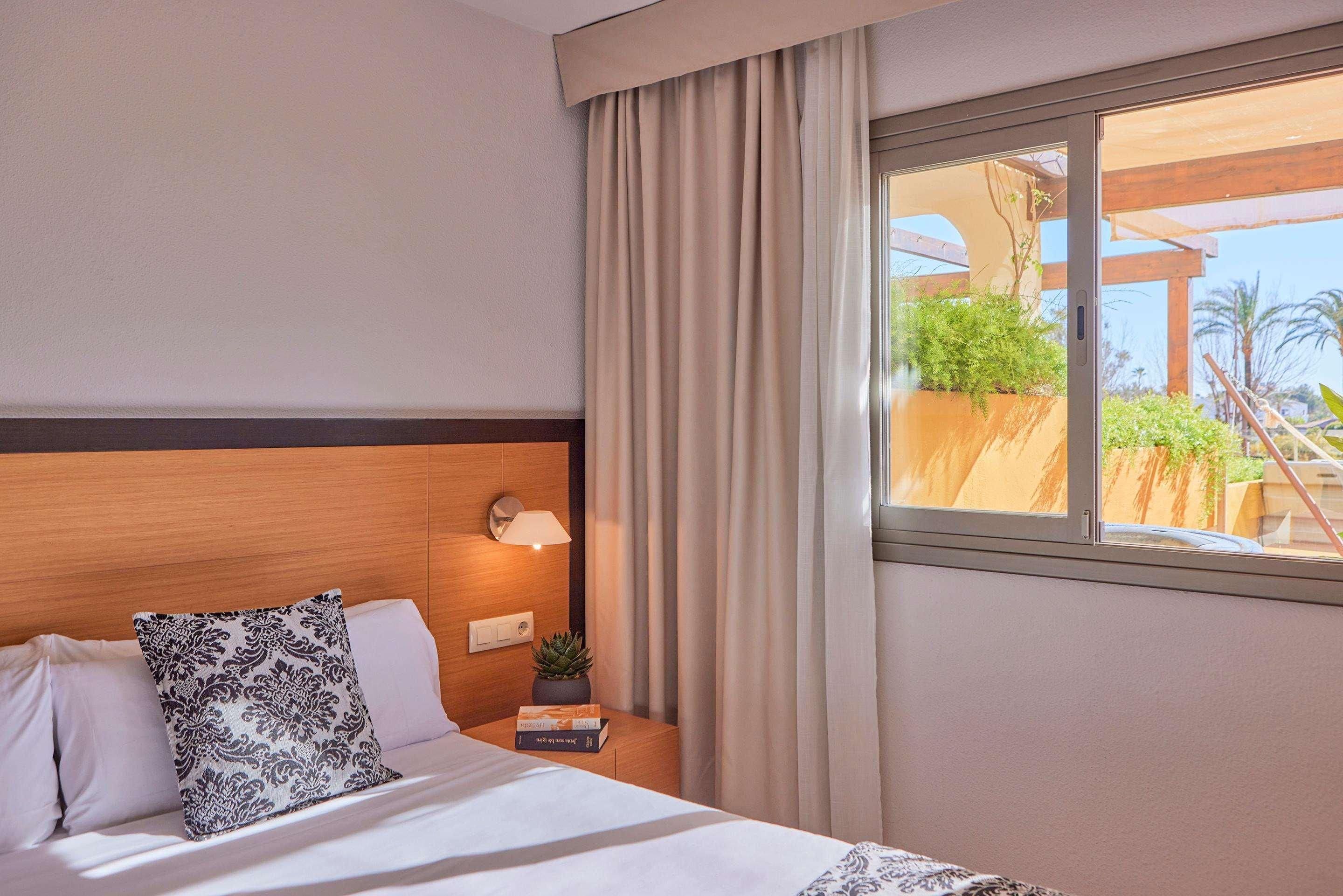 Habitación de hotel con cama, mesita de noche con lámpara, ventana con cortinas y vista a zonas verdes.