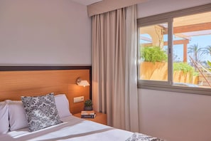 A hotel room with a bed, a nightstand with a lamp, a window with curtains, and a view of greenery outside. - Zafiro Tropic (Alcúdia)