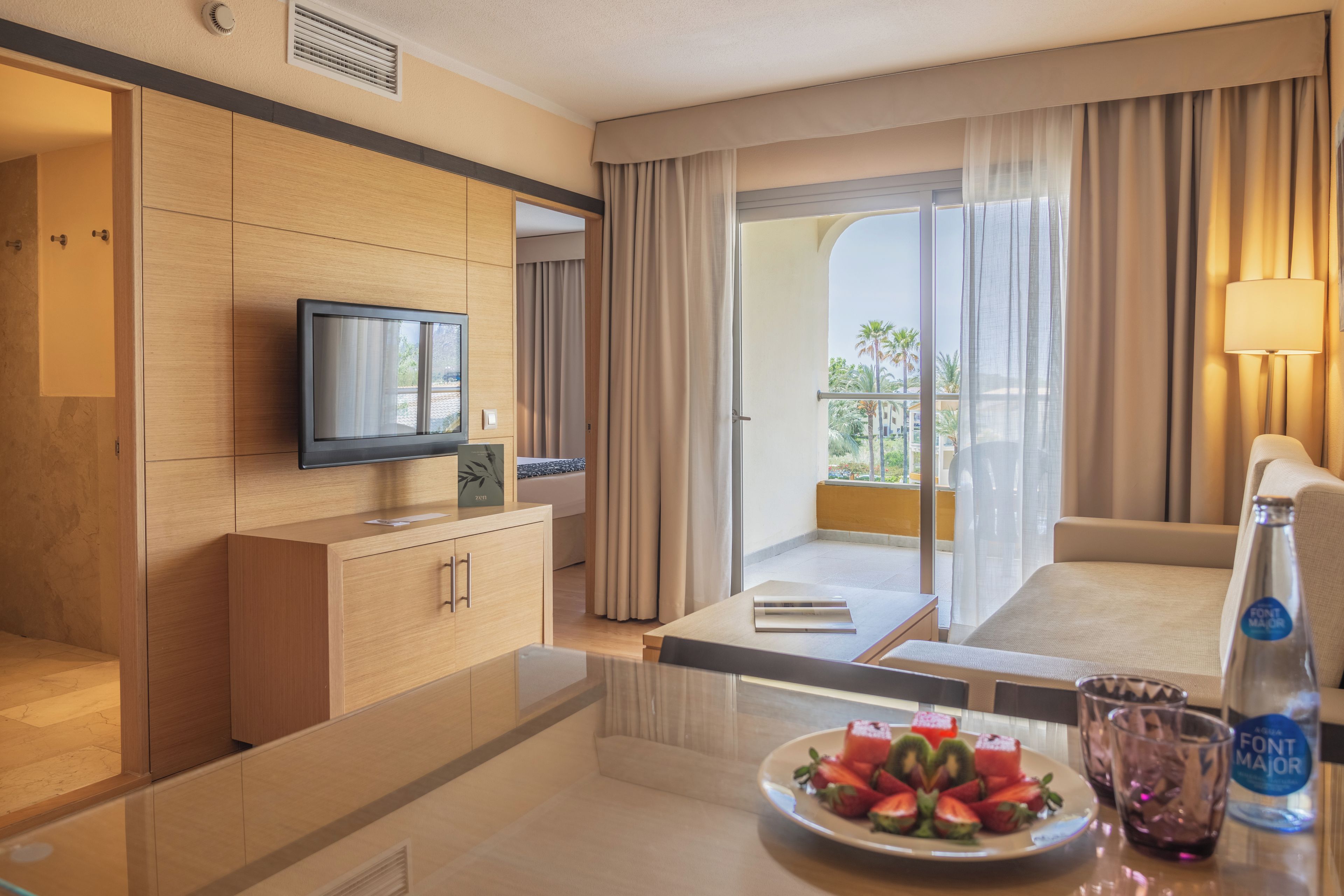 Een hotelkamer met een televisie, een eettafel met een schaal fruit, een bank en een balkon met gordijnen.