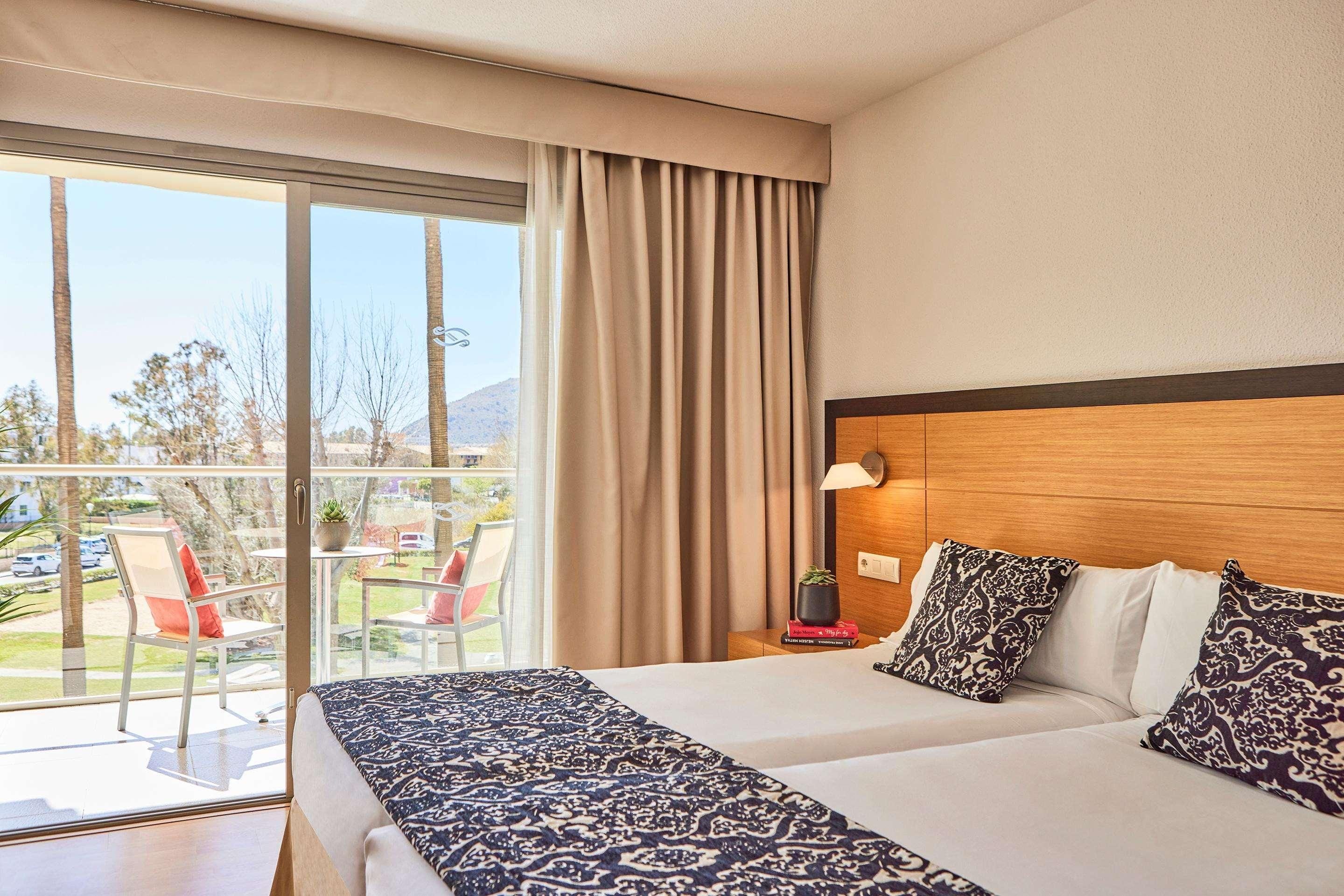 Habitación de hotel con una cama grande, cabecera de madera, mesita de noche con lámpara y vistas al exterior a través de una puerta corrediza de vidrio.