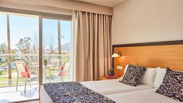 Habitación de hotel con una cama grande, cabecera de madera, mesita de noche con lámpara y vistas al exterior a través de una puerta corrediza de vidrio.