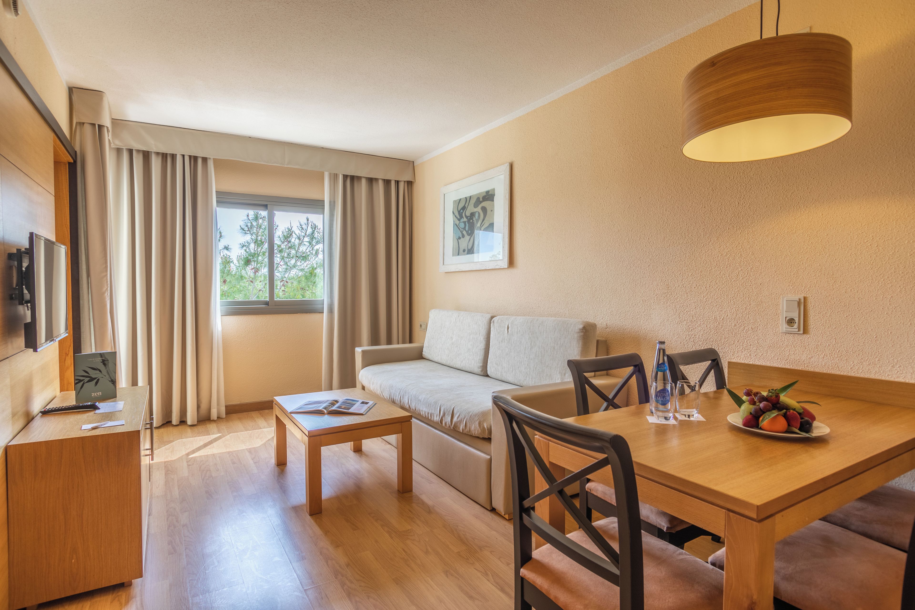 Habitación de hotel moderna con una mesa de comedor de madera, un sofá, una pequeña mesa de centro, un televisor y una ventana con cortinas.