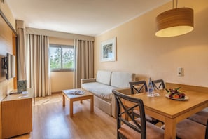 A modern hotel room with a wooden dining table, a sofa, a small coffee table, a TV, and a window with curtains. - Zafiro Tropic (Alcúdia)