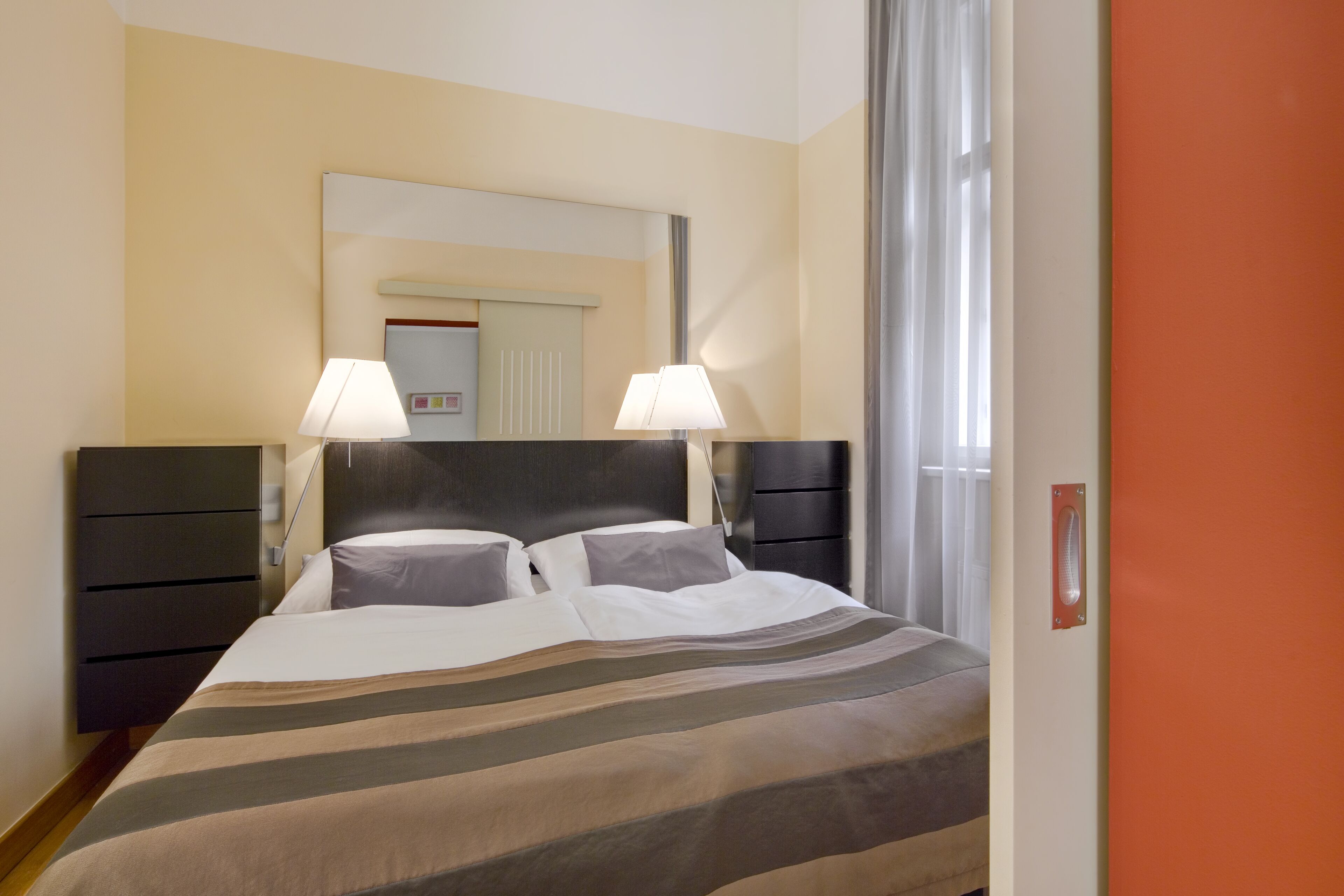 A hotel room with a bed, two bedside lamps, a nightstand, a mirror, and a window with curtains.