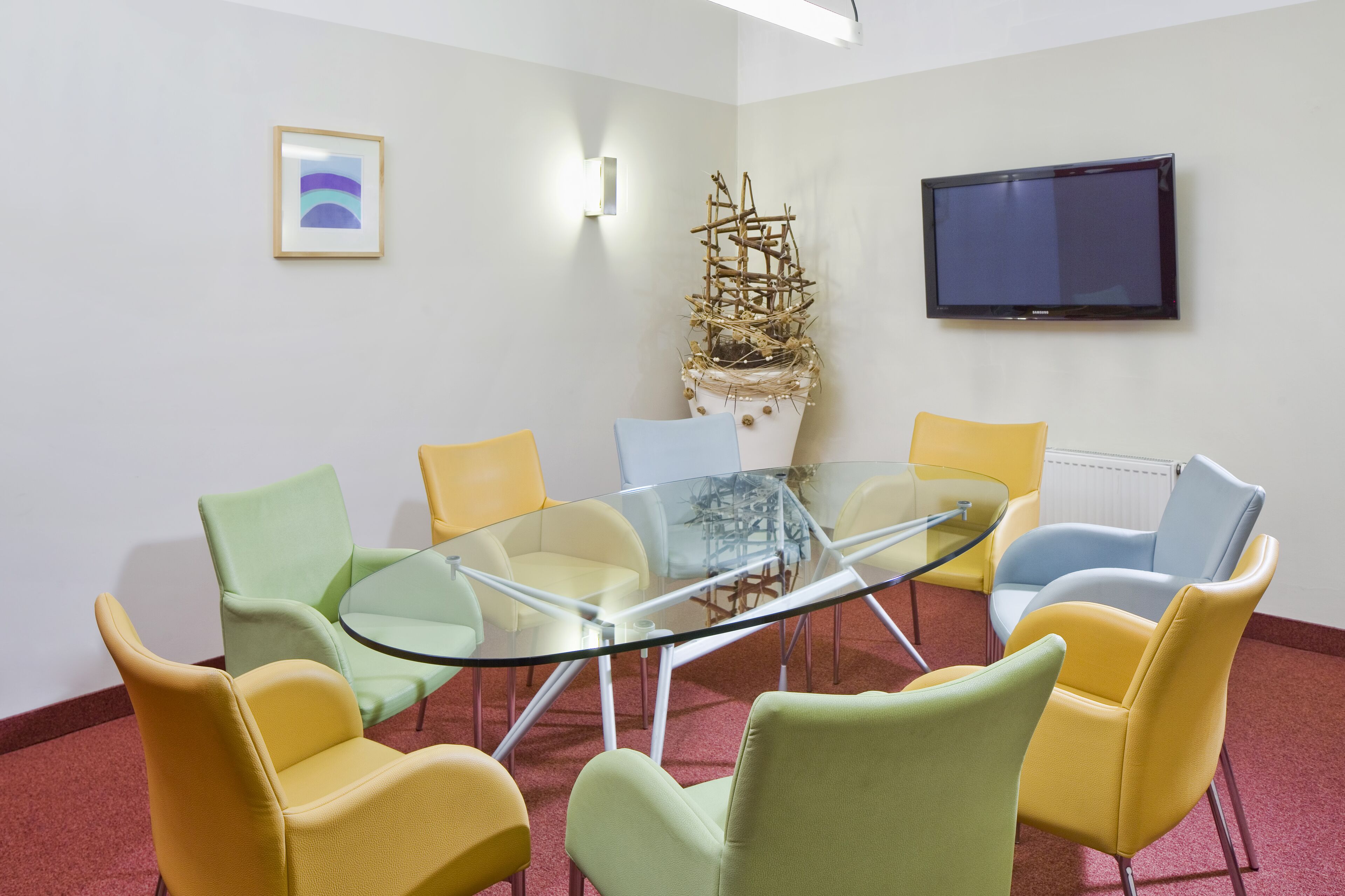 Un salón de conferencias con una mesa de vidrio rodeada de sillas coloridas, un televisor montado y una escultura decorativa.