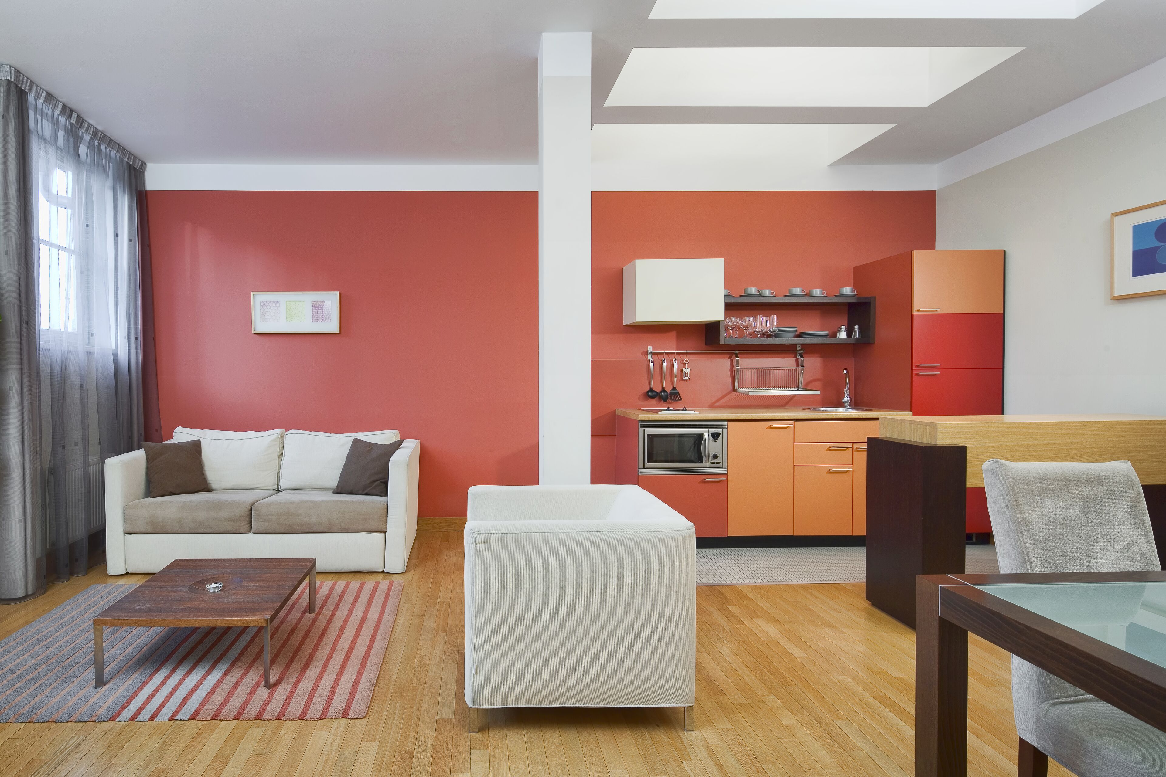 A modern living room with a sofa, ottoman, and coffee table, a kitchen with orange cabinets and a microwave, and a dining area with a table and chairs.