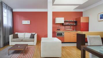 A modern living room with a sofa, ottoman, and coffee table, a kitchen with orange cabinets and a microwave, and a dining area with a table and chairs.