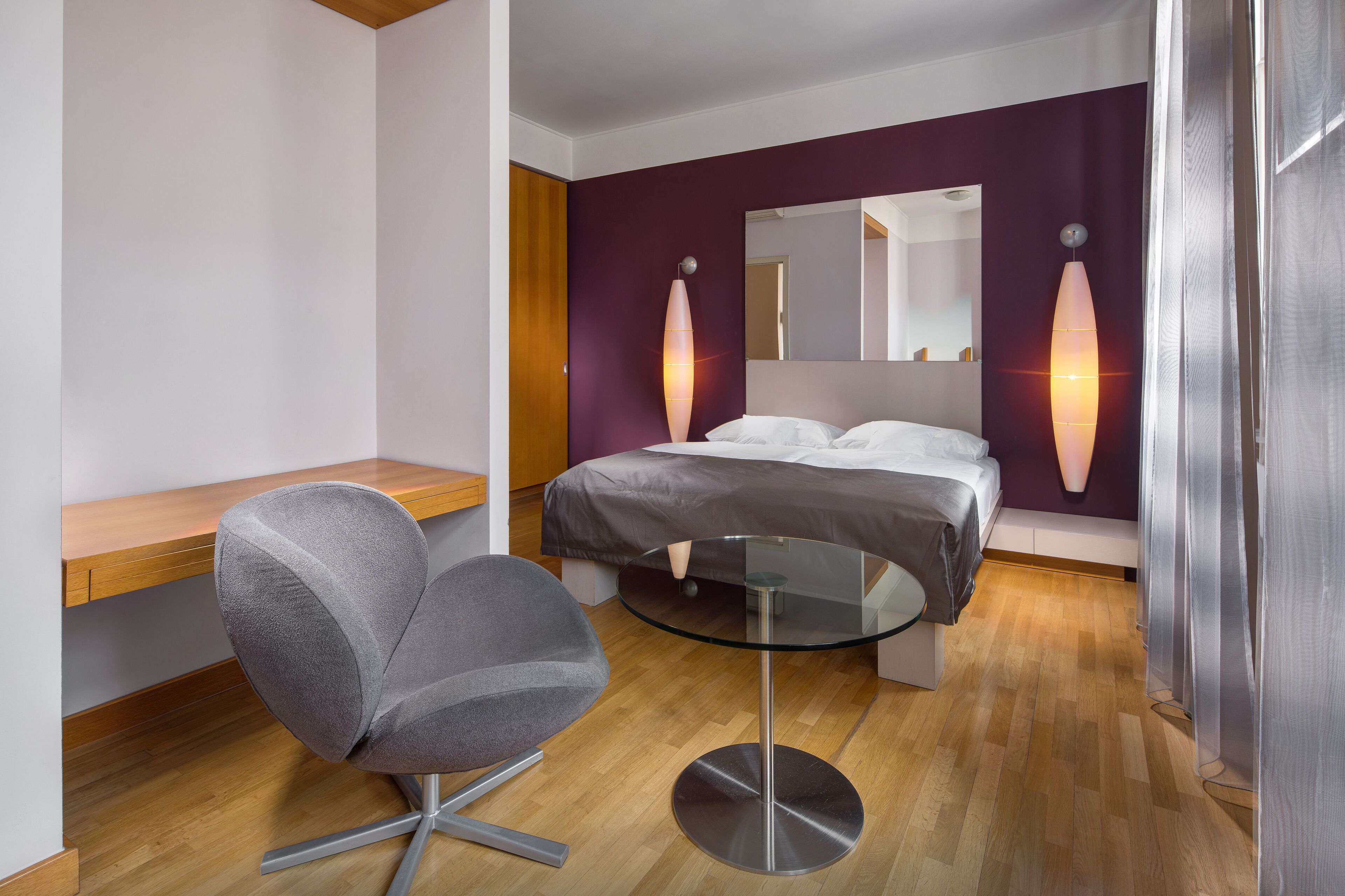 Ein modernes Hotelzimmer mit einem Holztisch, einem runden Glastisch, einem grauen Sessel, einem Bett mit weißer Bettwäsche und einem großen Spiegel.