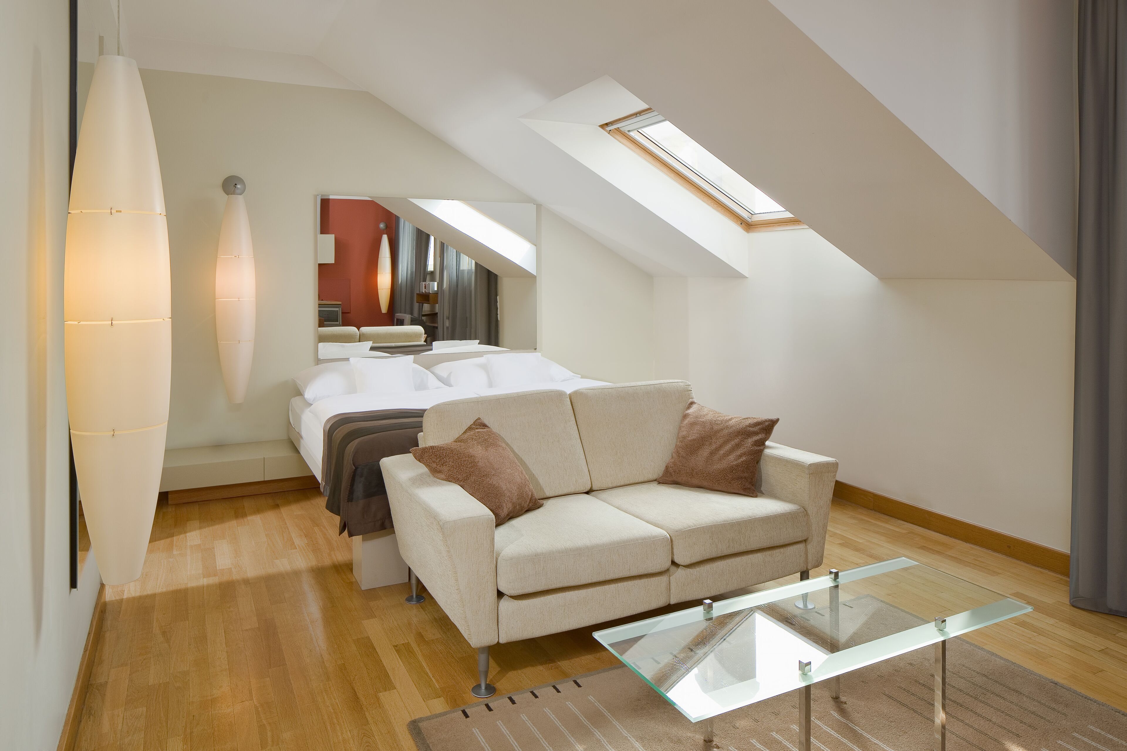 A modern loft bedroom with a sloped ceiling, a glass coffee table, a beige sofa, and a large mirror.