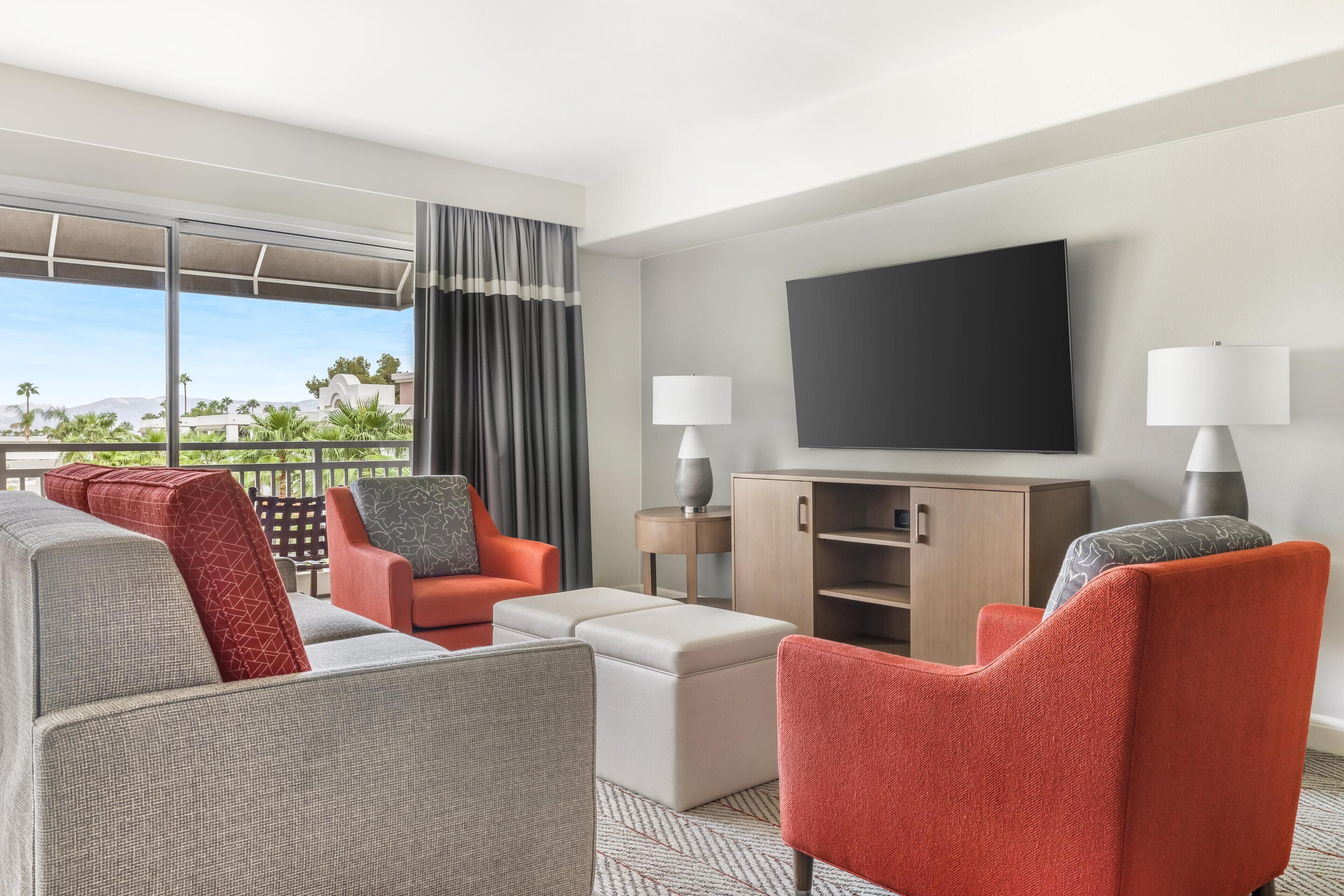A hotel room with a large window, a flat-screen TV, a sofa, a chair, a small table, and a cabinet.