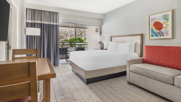 A hotel room with a bed, a sofa, a table, and a chair. There is a balcony with a view of greenery.