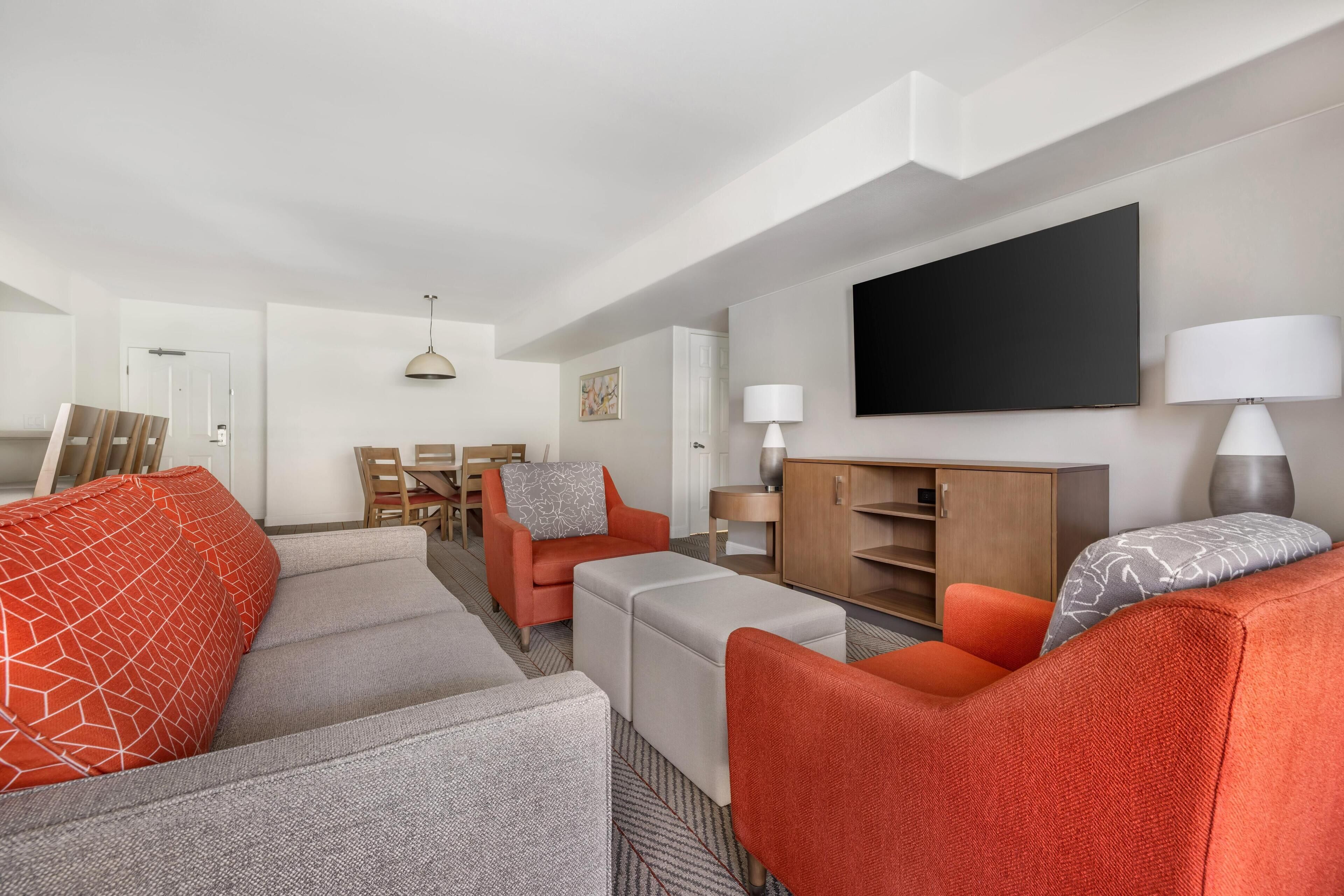 A modern living room with a sofa set, a dining table, and a flat-screen TV mounted on the wall.