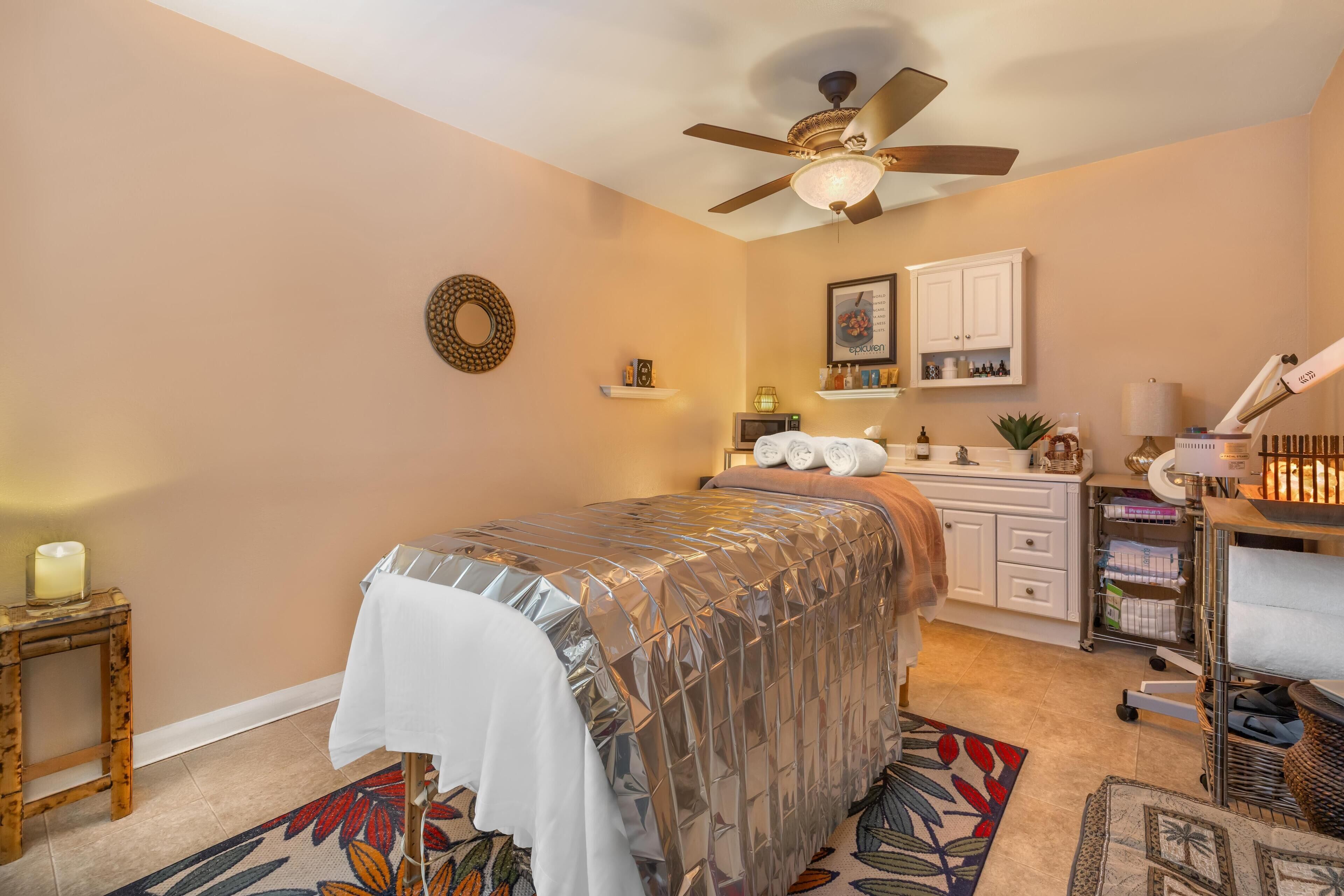 A bedroom with a bed, a ceiling fan, a small table with a candle, a wall-mounted shelf with toiletries, and a bathroom vanity with a towel rack.