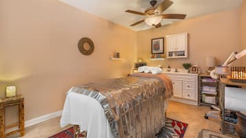 A bedroom with a bed, a ceiling fan, a small table with a candle, a wall-mounted shelf with toiletries, and a bathroom vanity with a towel rack.